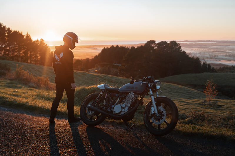man looking down at his motorcycle on a hill while the sun is setting in the background
